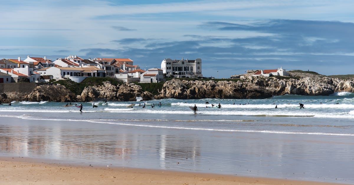 Surfers riding waves near a charming beach town with rocky shores.
