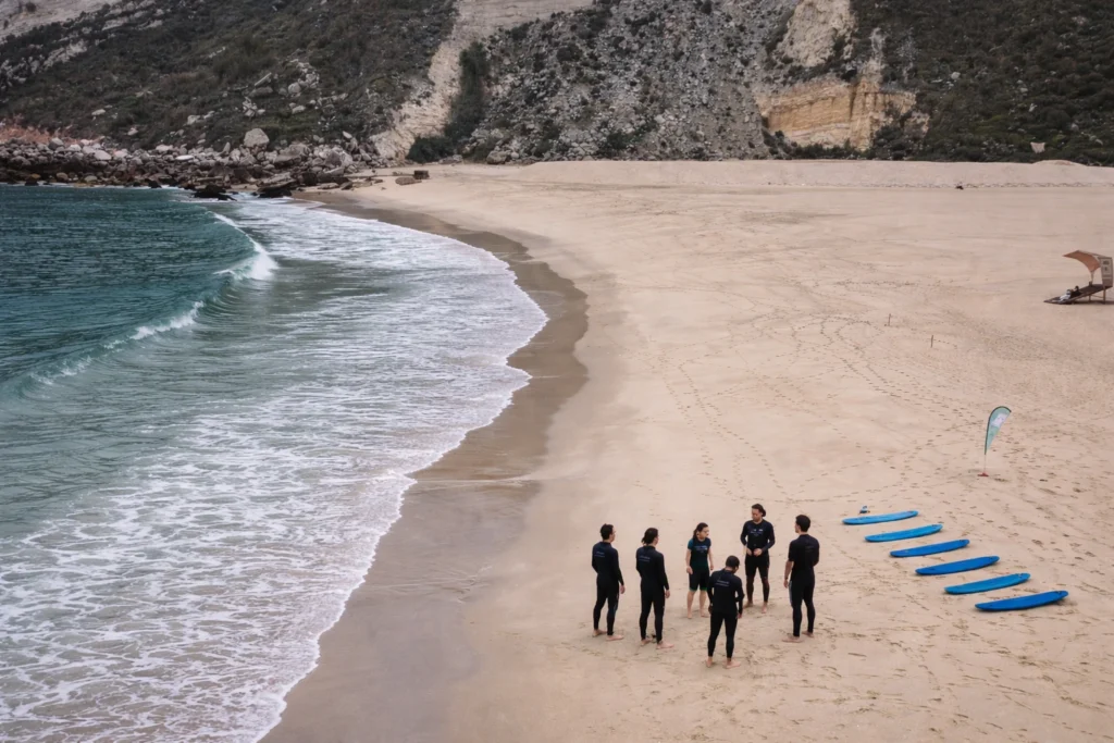 Aerial view of a surf lesson in Nazaré with students in wetsuits standing on the beach beside blue surfboards near the shoreline.
